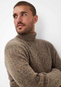 Brauner, gestrickter Rollkragenpullover mit einem strukturierten Muster, das blaue Sprenkel aufweist, und einem gerippten Kragen, vor einem neutralen Hintergrund gezeigt.