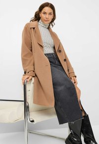 Brun uldblandet frakke med store knapper, parret med en mønstret turtle-neck og en mørk denimnederdel. Sorte knæhøje støvler fuldender looket.