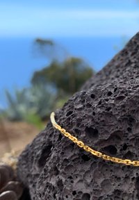 Goldfarbener Metallkettearmband mit kunstvollen Gliedern, auf einem strukturierten schwarzen Vulkangestein-Hintergrund, vor einem blauen Himmelshorizont.