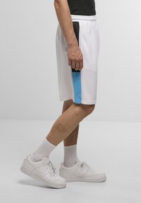 I pantaloni sportivi bianchi presentano una striscia laterale blu e dettagli neri, realizzati in un tessuto morbido. Abbinati a sneakers bianche e calze a costine.