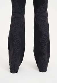 Schwarze Denim-Hosen mit aufwendiger, tonal gestickter Borte an den Seiten, die florale und abstrakte Muster darstellen, in einer taillierten Silhouette.