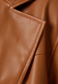 Detail hnědé umělé kožené bundy, se hladkým povrchem, ostrým klopovým límcem a čistým šitím. Pevný materiál s jemným leskem.