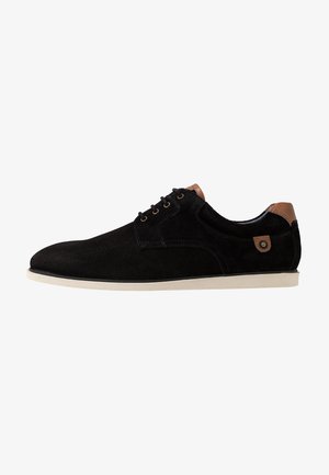 Chaussure noire en daim à lacets avec un bout arrondi, accent de talon beige contrastant, œillets en métal et semelle en caoutchouc blanche.