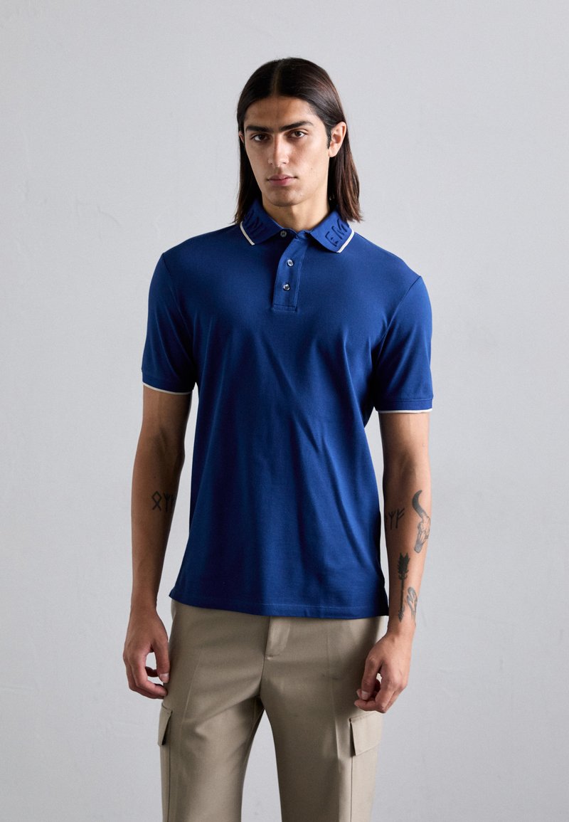 Bărbat cu păr lung și închis la culoare, îmbrăcat cu un polo blue cu mâneci scurte și pantaloni cargo bej, stând cu spatele la un fundal deschis.
