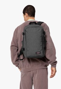 Grå tygryggsäck med rektangulär form, framdragkedja, sidspännen och handtag upptill. Justerbara remmar är fästa för bärande.