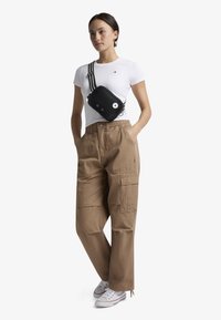 T-shirt bianco aderente, pantaloni cargo color khaki con tasche, borsa a tracolla nera con dettagli bianchi, tracolla a strisce bianche e nere, sneakers casual.