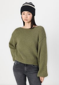 Pull en maille vert olive avec de larges manches et un ourlet côtelé, associé à un jean en denim noir et un bonnet bleu marine avec des rayures blanches.