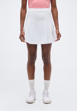 Inimene, kes kannab valget tennisseelikut, valgeid sokke, valgeid spordijalatseid ja roosat varrukateta särki, seisab ühevärvilise tausta ees.