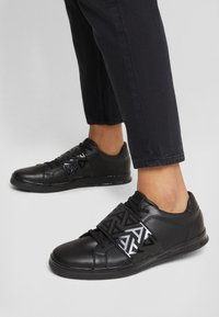 Svarta lädersneakers med geometriskt mönster i svart och silver, snörningsdesign och slät yta, som bärs med mörka smala byxor.