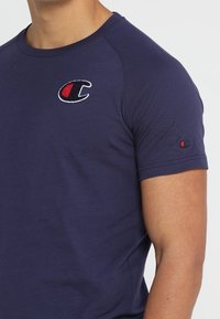 Muž v námořnické modré polokošili s krátkým rukávem, na prsou a levém rukávu s červeným a černým logem Champion.