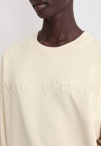 Crèmekleurige sweatshirt met een ronde halslijn, voorzien van een geborduurd "Marc O'Polo" logo in bijpassende draad. Zachte, gladde textuur.