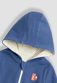 Modrá bunda s kapucňou zo štruktúrovanej látky, s krémovým podšívkou a malým vyšívaným detailom líšky na hrudi.