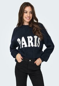 Tumed sinise kudumisega jumpers, millel on valged pehmed tähed sõnaga "PARIS". Pikkade varrukatega ja mugava lõikega. Mustad teksad täiendavad komplekti.