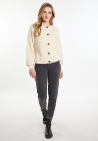Cardigan lavorato a maglia color crema con sei bottoni scuri, texture a costine, scollatura rotonda e vestibilità comoda. Indossato sopra pantaloni grigi e stivali neri.