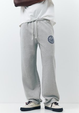 Bărbat purtând pantaloni de trening gri deschis cu un emblema albastru pe coapsa stângă și adidași albi cu tălpi negre.