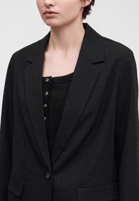 Personne portant un blazer noir sur un top noir côtelé avec des boutons, montrée des épaules au haut de la poitrine sur un fond clair.