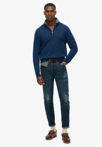 Suéter de punto azul marino con cuello con cremallera sobre una camisa de cuadros, combinado con jeans oscuros y zapatos de cuero marrón con cordones y calcetines.