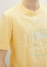 Camiseta de algodón amarilla con cuello redondo, que presenta texto en blanco y verde menta, y un patrón texturizado tipo slub.