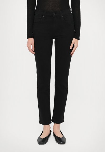Schwarze Skinny-Jeans mit hoher Taille, ausgestattet mit einem Reißverschluss und vorderen Taschen. Kombiniert mit schwarzen Ballerinas wirkt der Stoff glatt und elastisch.