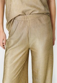 Złote metaliczne spodnie o szerokich nogawkach, z pionowym żebrowanym wzorem, elastycznym pasem i bocznymi kieszeniami. Materiał subtelnie błyszczy w świetle.