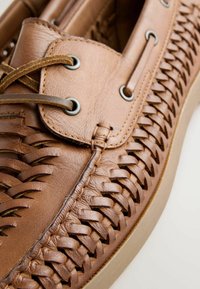 Chaussure en cuir marron avec motif tissé, devant à lacets, et semelle beige, montrée en gros plan.