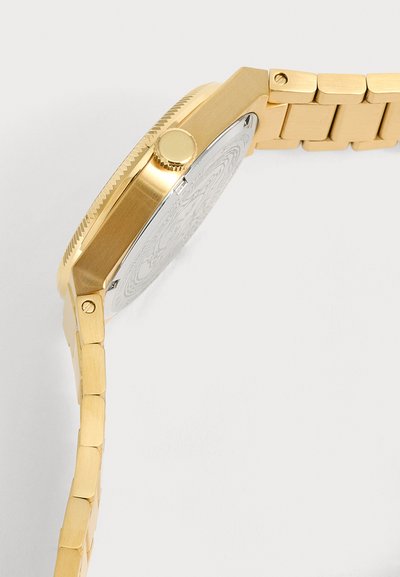 Montre-bracelet en métal doré avec bracelet à maillons et dos argenté à motifs, montrant le profil latéral et la couronne sur fond blanc.