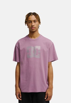 Młody mężczyzna z krótkimi warkoczykami, ubrany w luźny mauve t-shirt DC z dużym srebrnym logo, stojący na tle jasnego, jednolitego tła.