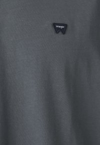 Tumši pelēks T-krekls, kas izgatavots no mīksta auduma, ar mazu melnu izšūtu Wrangler logotipu kreisajā pusē. Klasiska apaļa kakla dizains.