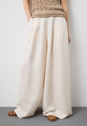 Mujer con pantalones anchos color crema, top beige de crochet y sandalias marrones de punta abierta, de pie contra una pared lisa de color gris claro.