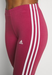 Tajta rosa träningsleggings med tre vertikala vita ränder och en liten vit Adidas-logotyp på den nedre vänstra sidan.