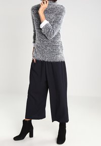 Maglione grigio fluffoso con maniche lunghe, abbinato a pantaloni neri a gamba larga. Stivali neri alla caviglia completano l'outfit. Design minimalista.