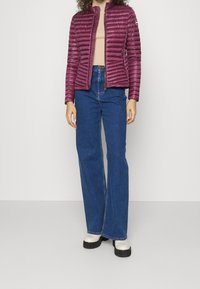 Lila puffjacka med quiltad design, i kombination med högmidjade blå jeans. Modell som bär vita plattformstövlar. Enkelt bakgrund.