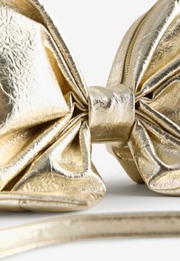 Kultainen keinonahkainen laukku, joka on muotoiltu suureksi rusetiksi. Siinä on teksturoitu pinta, keskellä vetoketjukiinnitys ja sileä olkahihna.