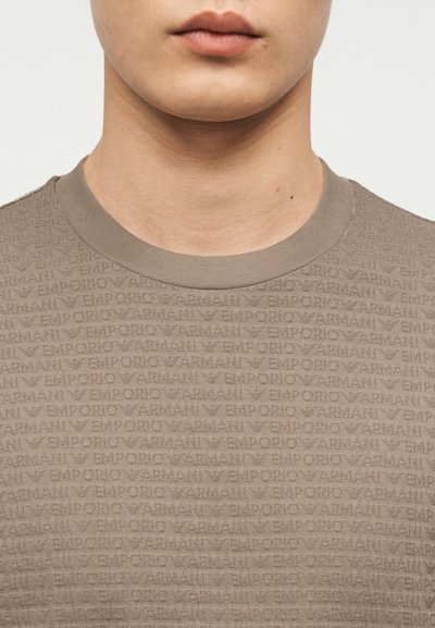 Κάτω μέρος του προσώπου και ο λαιμός ενός ατόμου που φορά ένα καφέ πουκάμισο Emporio Armani με ανάγλυφο λογότυπο και ριμπ λαιμόκοψη.