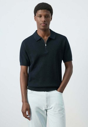 Joven de pie con la mano en el bolsillo, vestido con una camiseta polo negra de manga corta con cremallera y pantalones de color claro sobre un fondo liso.