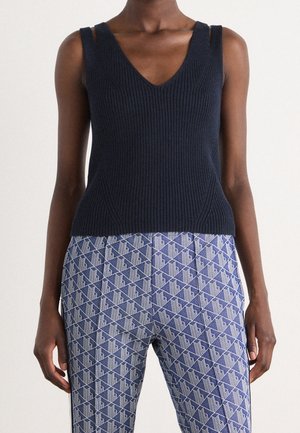 Femme portant un haut noir côtelé sans manches et un pantalon bleu taille haute avec un motif géométrique blanc, se tenant devant un fond uni.