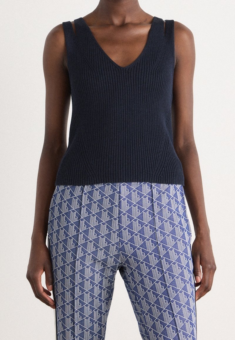 Femme portant un haut noir côtelé sans manches et un pantalon bleu taille haute avec un motif géométrique blanc, se tenant devant un fond uni.