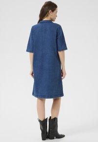 Denim jurk in donkerblauw, met korte mouwen en een kraag, met een rechte snit en zichtbare stiksels. Zwarte cowboylaarzen worden gedragen.