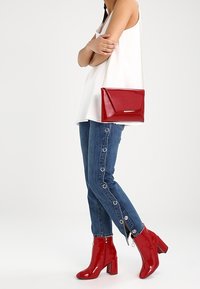 Femme portant un jean bleu avec des détails en anneaux métalliques, des bottines rouges en cuir verni, un haut blanc sans manches, et tenant un sac rouge avec une bandoulière chaîne.