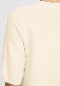 Kootud lühikeste varrukatega topp helesbeezis, millel on tekstuuriga kanga horisontaaltriibud, ümar kaelus ja ribatud ääreotsad.