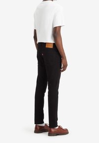 Jeans de mezclilla negra con un parche de cuero marrón en la parte trasera, combinados con una camiseta blanca y zapatos marrones, vistos desde la parte de atrás.