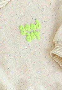 Sudadera beige claro con motas de colores. Presenta letras verdes en relieve que dicen "buen día" en una fuente divertida. Puños y cuello acanalados.