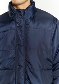 Giubbotto imbottito blu navy con colletto alto, tessuto liscio e lucido, chiusura con bottoni a pressione e texture a righe orizzontali. Design leggero.