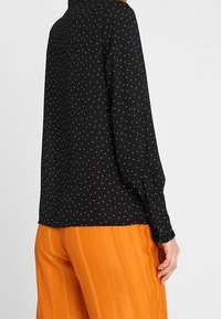 Schwarze Bluse mit langen Ärmeln und weißen Polka-Dots, mit einem knöpfbaren Bündchen und lockerer Passform. Kombiniert mit leuchtend orangefarbenen, plissierten Hosen.