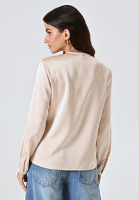 Blusa di raso color crema con maniche lunghe, leggero lucido e dettaglio a bottoni sul retro. Abbinata a pantaloni in denim blu chiaro.