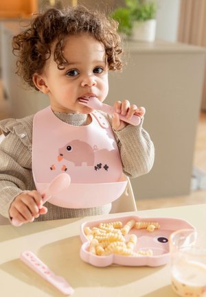 Peuter met krullend haar die een roze slabbetje draagt, een roze vork en lepel vasthoudt, zittend aan een tafel met pasta op een roze dierenbord.