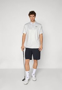 Krátký rukáv polokošile ve světle šedé barvě s jemným vzorem, černým logem Nike, v kombinaci s tmavě modrými šortkami a bílými teniskami. Drží golfovou hůl.