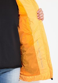 Leichte Jacke mit orangefarbenem Nyloninnenfutter und schwarzem Außenmaterial, ausgestattet mit einem Reißverschluss, glatter Textur und Pflegehinweisen im Inneren.