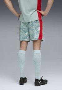 Pantaloni sportivi verde pallido con motivo astratto, pannello laterale rosso, calze azzurre alte fino al ginocchio coordinate, e scarpe nere con logo.