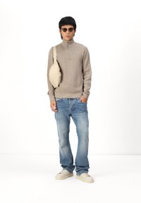 Beige ziptrui, lichtblauwe jeans met vervagingen, crèmekleurige sneakers en een beige crossbody tas. Model draagt ronde zonnebrillen.